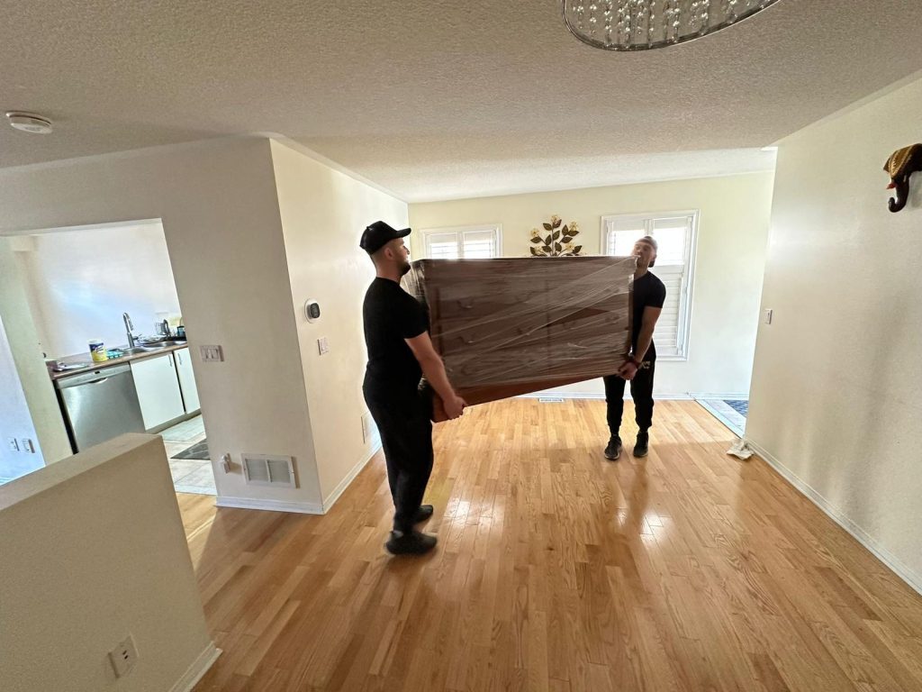 Two movers in black clothing carrying a large, plastic-wrapped item through a home.