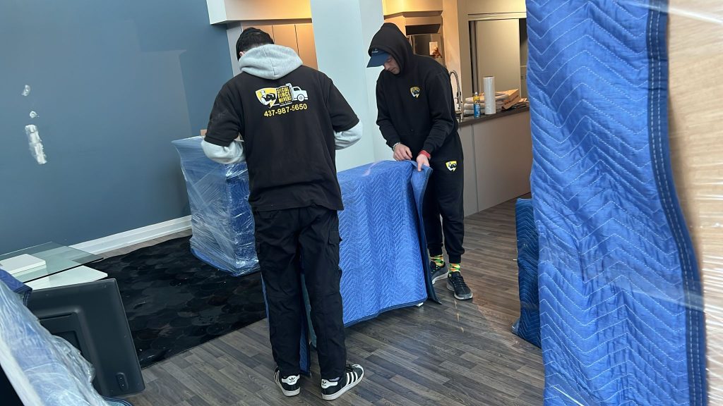 Two movers in branded Special Force Movers uniforms wrap furniture in protective blue moving blankets for a residential move